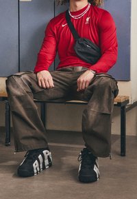 Rotes Langarmshirt mit Nike-Logo, braune Cargo-Hose, schwarze Umhängetasche und schwarze Sneaker mit weißen Akzenten, sitzend auf einer Bank.