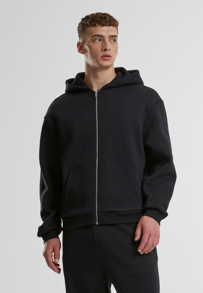 Hoodie zippé noir en tissu doux, avec une capuche spacieuse, des poches avant et des poignets côtelés, porté avec un pantalon assorti.