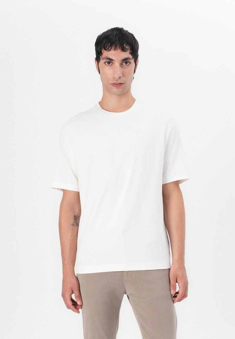 Tricou alb cu mânecă scurtă, cu o suprafață texturată, guler rotund și croială lejeră, asortat cu pantaloni khaki deschis.