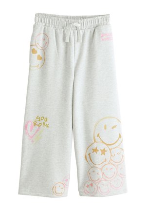 Barn med flettet hår har hvid cropped t-shirt, lysegul sweater omkring skuldrene, lysegrå joggingbukser med farverige smiley-ansigter og hvide sneakers.