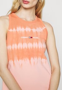 Top sin mangas de teñido anudado en naranja con un escote redondeado y costuras visibles. Presenta el logo "TOMMY JEANS" en blanco en la parte delantera. Textura de tela suave.