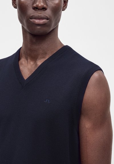 Gros plan sur une personne portant un pull sans manches bleu marine à col en V avec un petit logo brodé sur la poitrine.