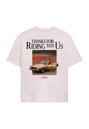 Giovane uomo che indossa una t-shirt oversize rosa chiaro, un cappellino da baseball verde scuro, una sciarpa coordinata al collo e pantaloncini verde scuro, che guarda di lato.