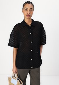 Chemise noire transparente à boutons avec un motif en losanges texturé, manches courtes et boutons argentés. Associée à un pantalon ample et un sac en osier.