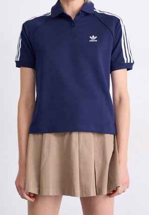 Osoba ubrana w granatową koszulkę polo Adidas z białymi paskami na rękawach i beżową plisowaną spódnicę na jednolitym tle.