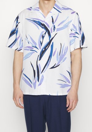 Mann trägt weißes Kurzarmhemd mit blauen und violetten abstrakten Blattmustern und dunkelblauer Hose, schaut nach vorne.