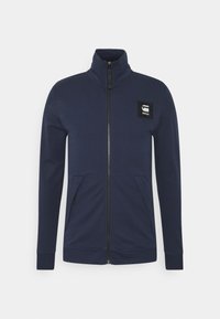 Marinblå zip-jacka med hög krage, två framfickor och en fyrkantig svart logotyp på bröstet, tillverkad av mjukt texturerat tyg.