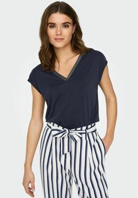 T-shirt bleu marine à col en V et manches courtes, avec un col texturé. Associe à un pantalon blanc, rayé verticalement en bleu marine, cintré à la taille.