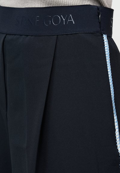 Marineblauwe geplooide broek met een gebrandmerkte tailleband en contrasterende lichtblauwe stiksel langs de zijkant. Gladde stoftextuur.