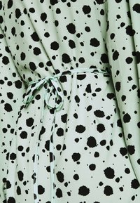 Tissu vert clair avec un motif éclaboussé noir, doté d'une ceinture assortie nouée à la taille. Texture lisse, avec des détails froncés.