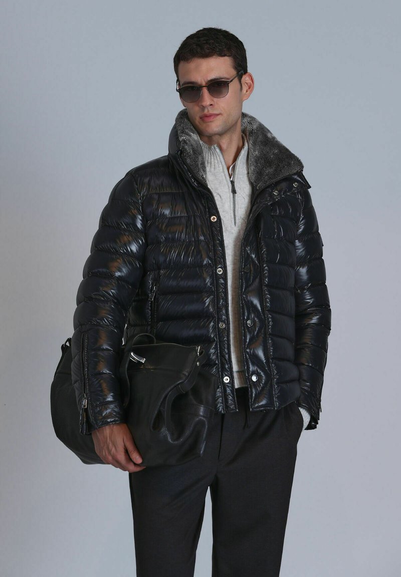 Schwarze Puffjacke mit glanzender Oberfläche, grauem Pelzkragen, mit Reißverschlusstaschen, getragen über einem hellgrauen Pullover, kombiniert mit schwarzen Hosen und Tasche.