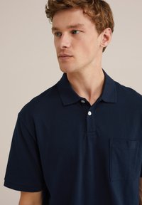 Polo majica temno modre barve iz bombaža, z ovratnikom, dvema gumboma in žepom na levi strani prsi z diskretnim logotipom. Gladka tekstura.