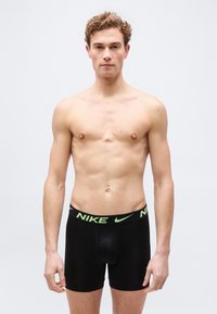 Νεαρός άνδρας με σγουρά μαλλιά φοράει μαύρα boxer σλιπ Nike, στέκεται μπροστά σε απλό φωτεινό φόντο, κοιτάζοντας προς τα εμπρός.