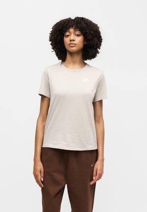 Vrouw met krullend haar, die een beige Nike T-shirt en een bruine Nike broek draagt, staand tegen een witte achtergrond.