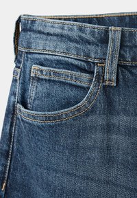 Gros plan sur un jean en denim bleu montrant la boucle de ceinture, la taille et la poche avant avec des détails de couture orange visibles.