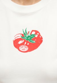 Λευκό πουκάμισο με κόκκινο γραφικό ντομάτας με πράσινα φύλλα και ένα μικρό κυκλικό αυτοκόλλητο με κείμενο πάνω στην ντομάτα.