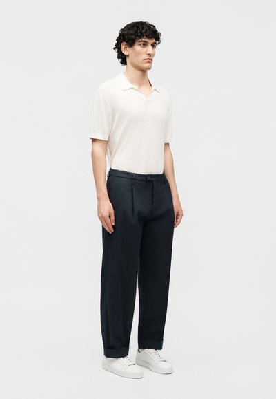 Jeune homme aux cheveux bouclés portant un polo blanc à manches courtes, un pantalon large foncé et des baskets blanches, debout devant un fond uni.