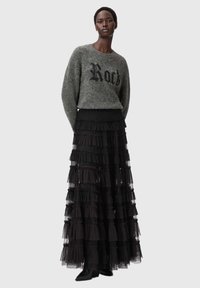 Nainen, joka pukeutunut harmaaseen pörröiseen neulepaitaan, jossa on teksti "Rock", yhdistettynä pitkään mustaan kerrokselliseen röyhelöhameeseen ja mustiin nilkkureihin, seisoo harmaalla taustalla.