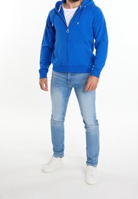 Haut à fermeture éclair bleu avec une poche kangourou, capuche ajustable avec cordon de serrage, associé à un jean en denim clair et des baskets blanches.