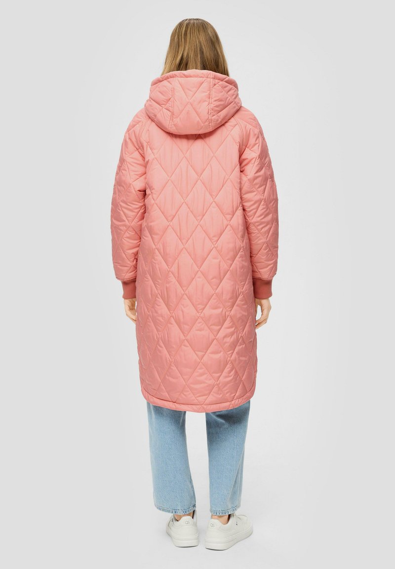 Langer rosa gesteppter Mantel mit Kapuze, versehen mit Rautenmustern, elastischen Bündchen und einer Länge bis zur Mitte der Oberschenkel, kombiniert mit hellblauen Jeans und Sneakers.