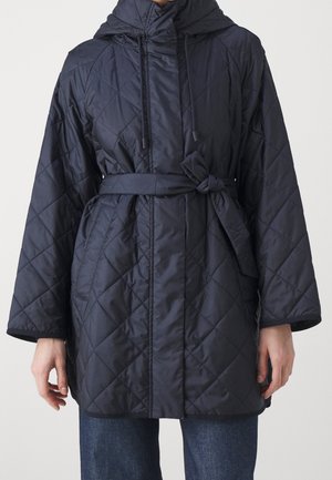 Vrouw draagt een marineblauwe doorgestikte jas met capuchon en een stoffen ceintuur om de taille, gecombineerd met een blauwe spijkerbroek.