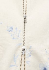 Κοντινή λήψη μπεζ υφάσματος με διπλό ασημί φερμουάρ και διακριτικά μπλε λουλουδάτα σχέδια και στις δύο πλευρές του φερμουάρ.