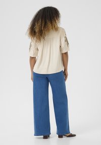 Chemise beige clair avec des détails brodés sur les manches, portée avec un pantalon en denim bleu à jambes larges. Les cheveux sont bouclés et volumineux, tombant au-delà des épaules.