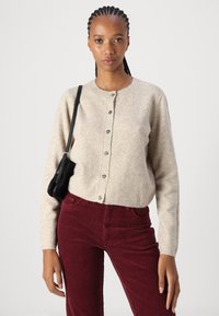 Beige ullcardigan med rund hals, knappfeste foran og ribbestrikkede mansjetter. Sammen med burgunder kordfløyelsbukser og en svart skulderveske.