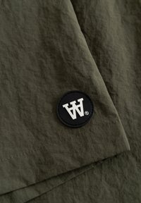 Tissu vert olive avec un patch circulaire noir présentant des lettres blanches stylisées en double 'A' près d'un ourlet cousu.