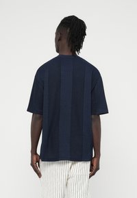 Homme à la peau foncée et aux cheveux tressés portant une chemise ample bleu marine à manches courtes et un pantalon blanc avec de fines rayures verticales noires, de dos.