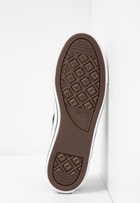 Semelle d'une basket blanche avec une semelle en caoutchouc marron présentant des motifs en losange et un logo étoile au centre, posée contre un mur d'angle blanc.