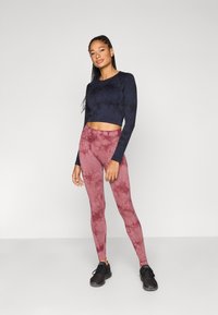 Μπλε ναυτικό crop top με μακριά μανίκια και ροζ κολάν με μοτίβο tie-dye, κατασκευασμένα από ελαστικό ύφασμα, συνδυασμένα με μαύρα αθλητικά παπούτσια.