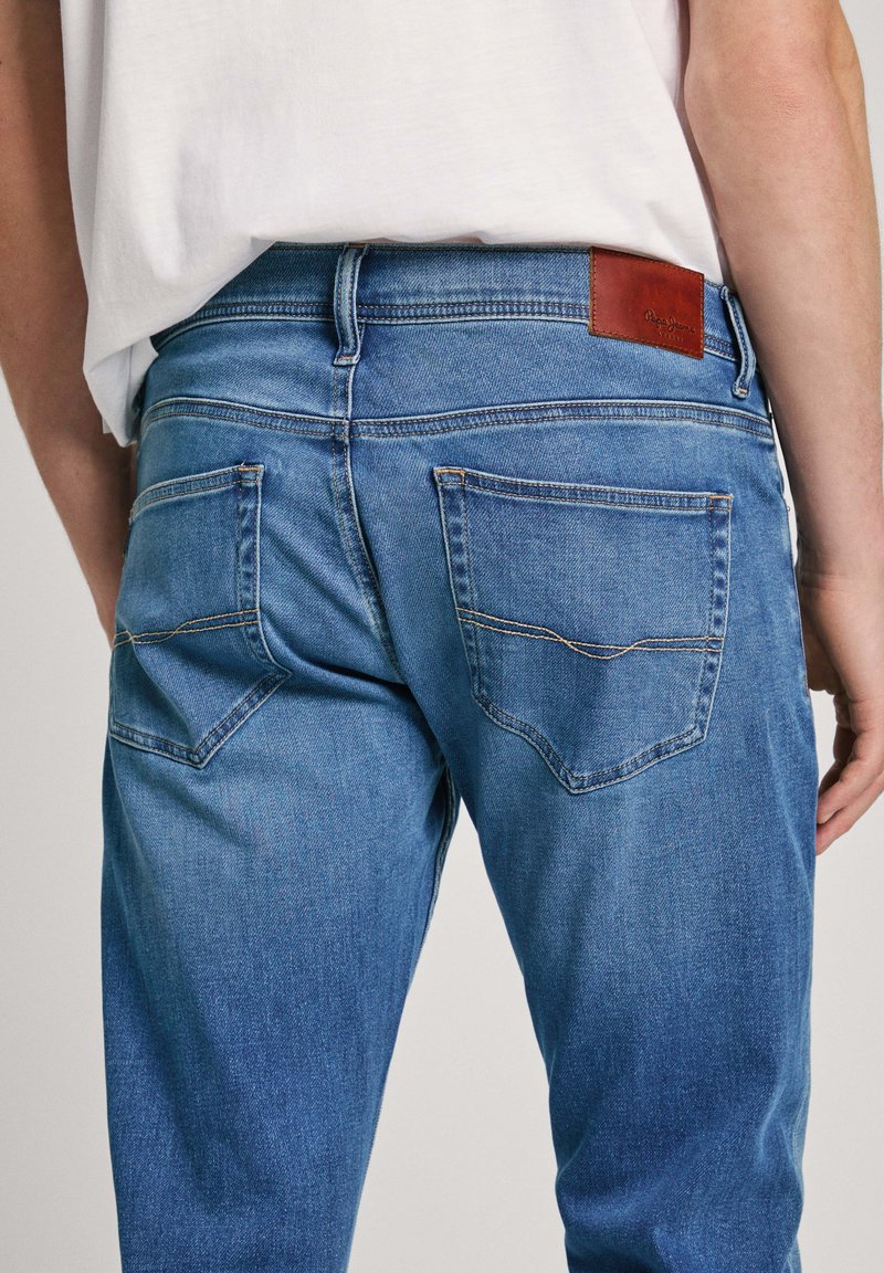 Jeans de mezclilla azul con un aspecto ligeramente desgastado, que presentan un parche de marca de cuero marrón y dos bolsillos traseros con un diseño de costura curvada.
