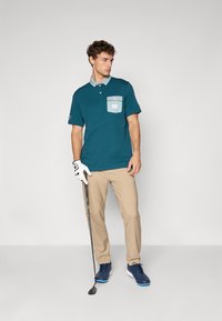 Mörk teal piké skjorte med lys grå krage og lomme, kombinert med khaki bukser og marineblå golf sko, som holder en golfklubbe.