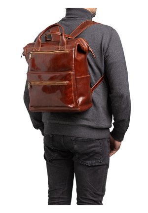 Man draagt een donkergrijze trui en jeans met een bruine leren rugzak met meerdere ritssluitingen en handvatten.