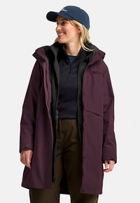 Giacca impermeabile di colore viola scuro con cappuccio removibile, tasche con cerniera e fodera interna nera, indossata sopra una maglia blu navy, abbinata a un berretto blu navy.