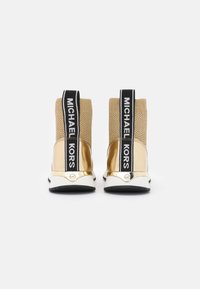 Złote i beżowe sneakersy z plecionym wierzchem, czarnym paskiem z logo oraz metalicznymi akcentami na pięcie, osadzone na białej podeszwie środkowej z czarną podeszwą zewnętrzną.