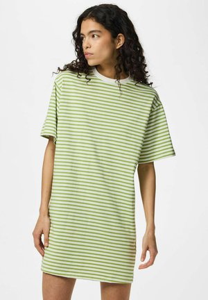 Kvinde med krøllet hår iført kort grøn og hvid stribet oversized t-shirt kjole, der kigger til siden mod en hvid baggrund.