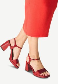 Röda lackläder sandaler med blockklack och öppen tådesign, samt ankelrem med spänne, mot en livlig bakgrund.