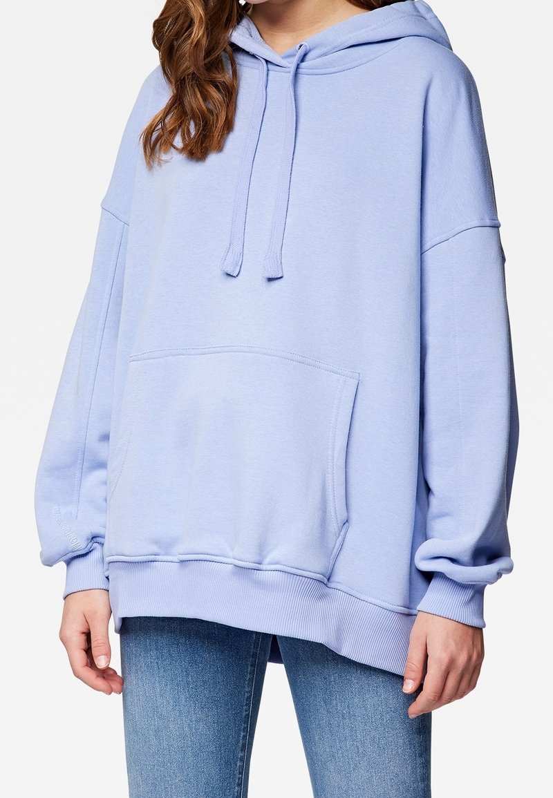 Sudadera azul claro extragrande con bolsillo frontal y cordones, usada con jeans azules, mostrada desde el pecho medio hasta las caderas.