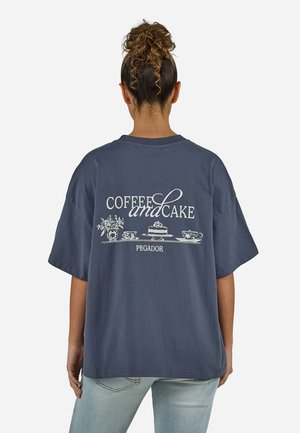 Kobieta z kręconym kokiem, ubrana w granatowy, oversize'owy T-shirt z białym napisem "Coffee and Cake" oraz ilustracjami ciasta, filiżanek i kwiatów na plecach.