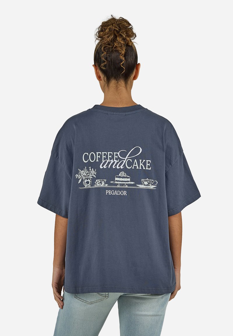 Γυναίκα με σγουρό κότσο φορώντας υπερμεγέθη μπλε ναυτικό T-shirt με λευκό κείμενο "Καφές και Κέικ" και εικονογραφήσεις κέικ, φλιτζανιών και λουλουδιών στην πλάτη.