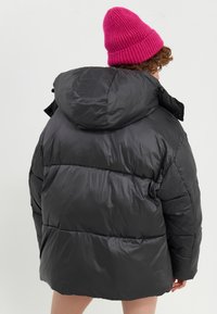 Svart boblejakke med hette, preget av en quiltet tekstur og synlige sømmer. Kombinert med en lys rosa strikket lue.