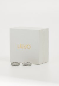 Strieborné kruhové náušnice s viacerými jasnými rhinestonmi, vystavené vedľa štruktúrovanej bielej krabičky s názvom značky "LIU·JO" v zlatej farbe.