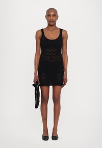 Rochie neagră tricotată, cu un design ajustat, decorată cu un model de găuri deschise. Însoțită de o geantă împletită asortată.