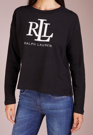 Kvinde iført sort langærmet skjorte med hvidt "RLL Ralph Lauren"-logo kombineret med blå jeans mod en neutral baggrund.