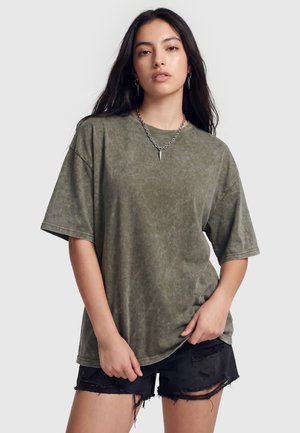 Ung kvinde med langt mørkt hår iført en oversized falmet grøn t-shirt, sorte revnede shorts og en spikes halskæde mod en ensfarvet baggrund.