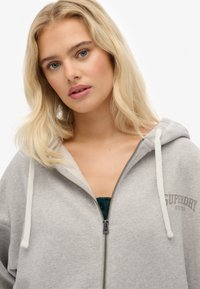 Sudadera con cremallera en color gris, con un bolsillo tipo canguro. Presenta cordones blancos, puños acanalados y un logo impreso en el pecho. Textura suave.