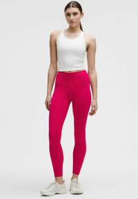 Bela majica na kratke rukave s tankimi naramnicami, v kombinaciji z visokimi, živimi roza leginami. Model nosi svetle športne čevlje. Gladek, raztegljiv material.