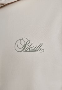 Kremno obarvan tekstil je okrašen z napisom »SikSilk« v zelenem pisavnem slogu. Površina je gladka z rahlim leskom.